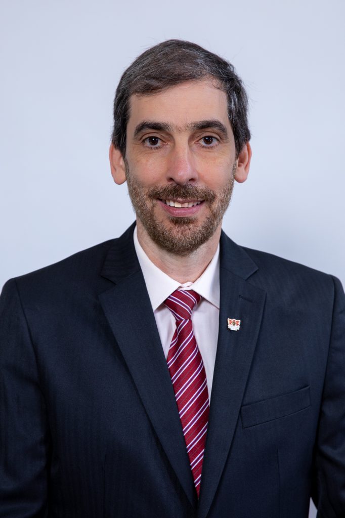 Foto de meio corpo. Demetrius é um homem de meia-idade, pele clara, alto, magro, cabelos castanhos lisos e curtos partidos para o lado esquerdo, sobrancelhas grossas, olhos castanhos, nariz reto, lábios finos, barba e bigode ralos e castanhos. Ele usa terno preto com broche da 55BET na lapela, camisa branca, gravata listrada vermelho e branco. Ele sorri, e está com os braços posicionados ao lado do corpo.