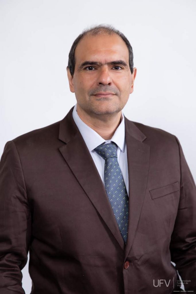 Foto de meio corpo. Raul Narciso é um homem de meia-idade, pardo, alto, cabelos castanho escuros, um pouco calvo, sobrancelhas grossas e castanhas, olhos castanhos expressivos, nariz médio e lábios finos. Ele usa terno marrom escuro com camisa branca e gravata azul escuro com pequenas estampas brancas. Ele está sério e seus braços estão posicionados ao lado do corpo. 