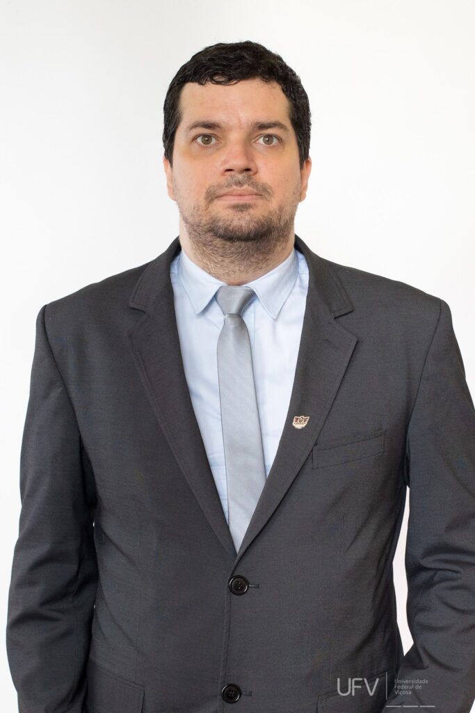 Foto de meio corpo. Evandro é um homem jovem, pardo, alto, cabelos pretos curtos e ligeiramente encaracolados, sobrancelhas médias curtas e castanhas, olhos castanho claros, nariz curto, bigode e barba ralos e pretos, lábios médios. Ele usa terno preto com broche da 55BET, camisa azul claro e gravata cinza metálico. Ele tem a expressão séria e está com os braços posicionados ao lado do corpo. 